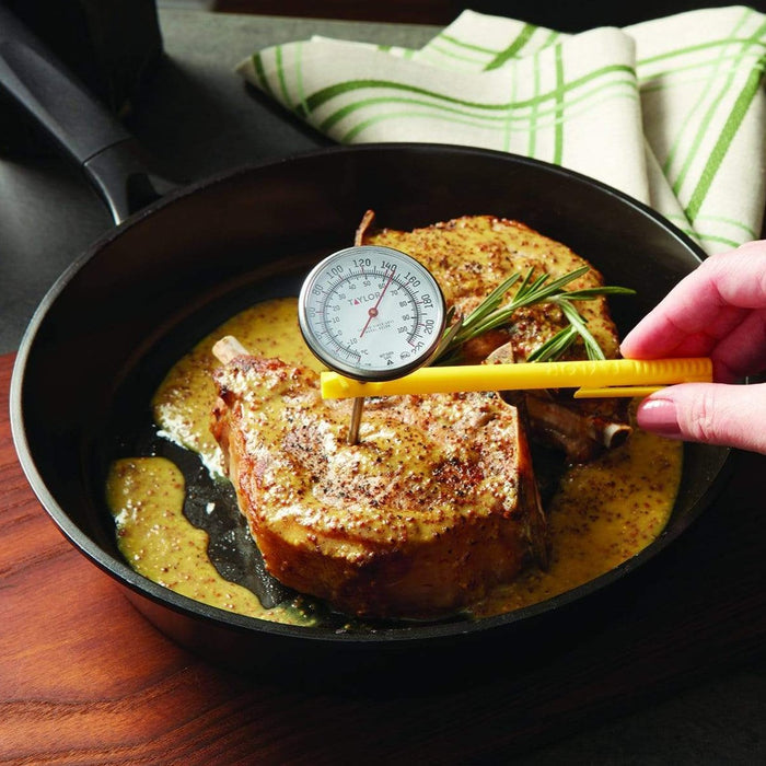 Hand holds a yellow instant read thermometer to check a seared pork chop in a skillet with rosemary.