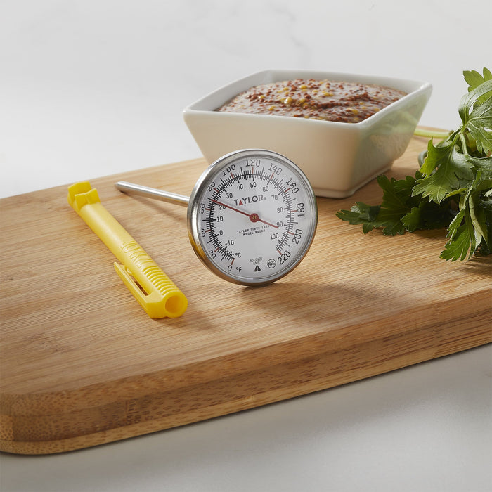 Taylor instant read thermometer with yellow handle and large dial placed on a wooden counter beside a white sauce dish.