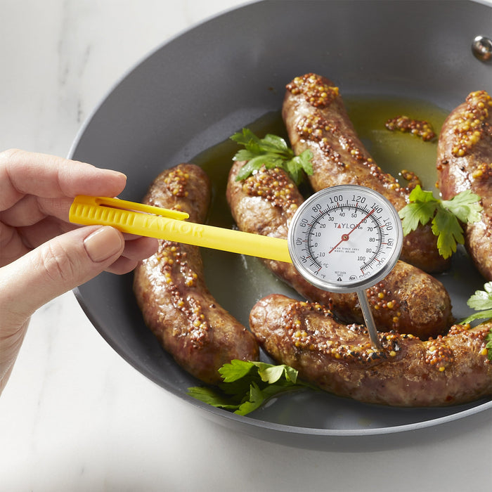 Close up of sausages coated with mustard seeds in a skillet being checked with a yellow handled instant read thermometer.