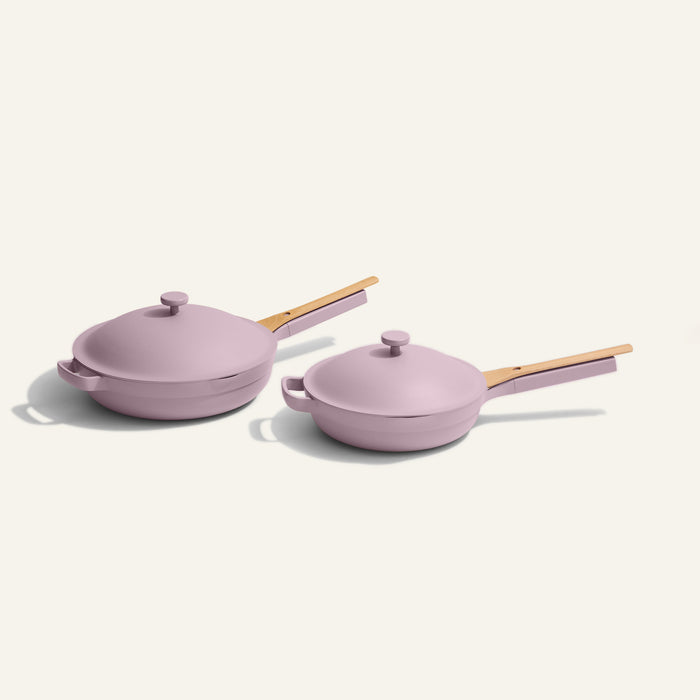 The Always Pan Duo, a set of two pink nonstick frying pans in different sizes with wooden handles and matching lids, offers versatile style and function for your kitchen, all displayed on a plain light background.