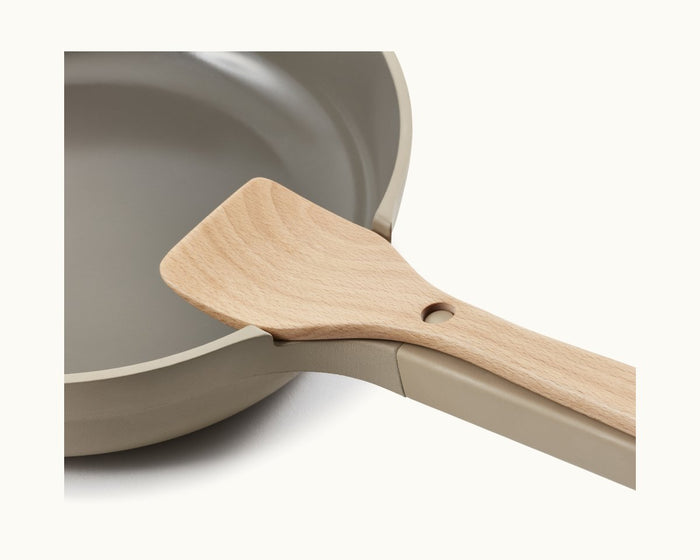 A close-up of a beige nonstick frying pan with a Beechwood Spatula—part of the Always Pan accessories—resting on its handle.