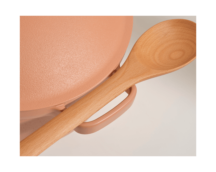 A close-up of a Beechwood Spoon resting on the handle of a pink pot with a textured lid, highlighting elegant kitchen utensils.