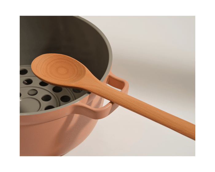 A Beechwood Spoon rests on the rim of a pink pot with a gray interior and metal steamer insert, making it a charming addition to your kitchen utensils.
