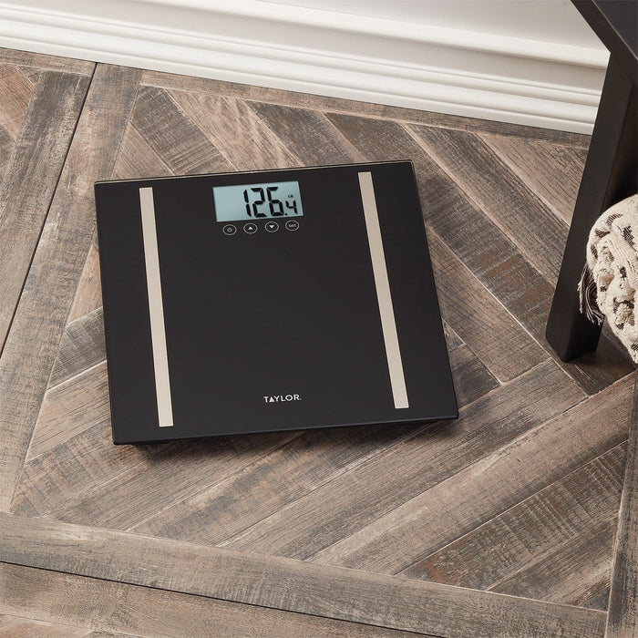Top-down view of Taylor black glass body composition scale on a wooden floor with display reading 126.4 lb