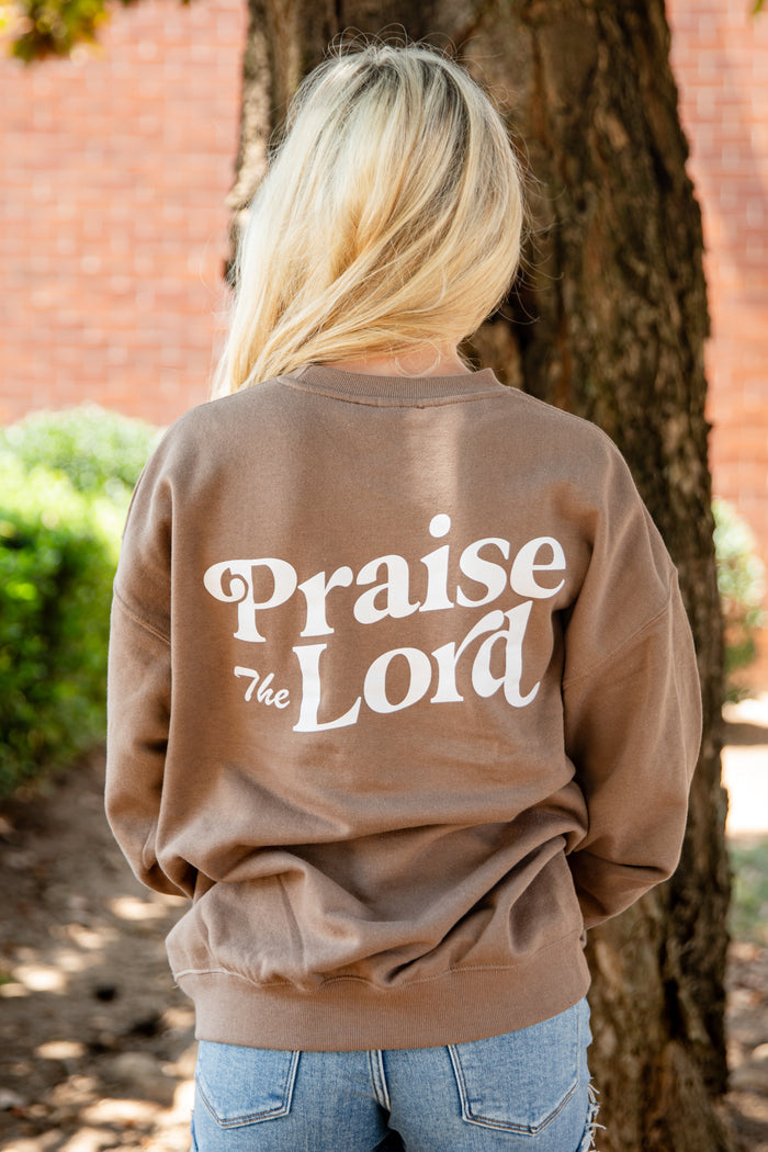 A woman viewed from behind wearing a brown crewneck sweatshirt with 'Praise The Lord' printed in white on the back, standing outdoors next to a tree.