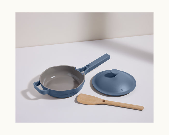 The Mini Always Pan 2.0 in blue, featuring a matching lid and wooden spatula, is displayed on a white surface—highlighting its versatile, nonstick design for easy cooking.