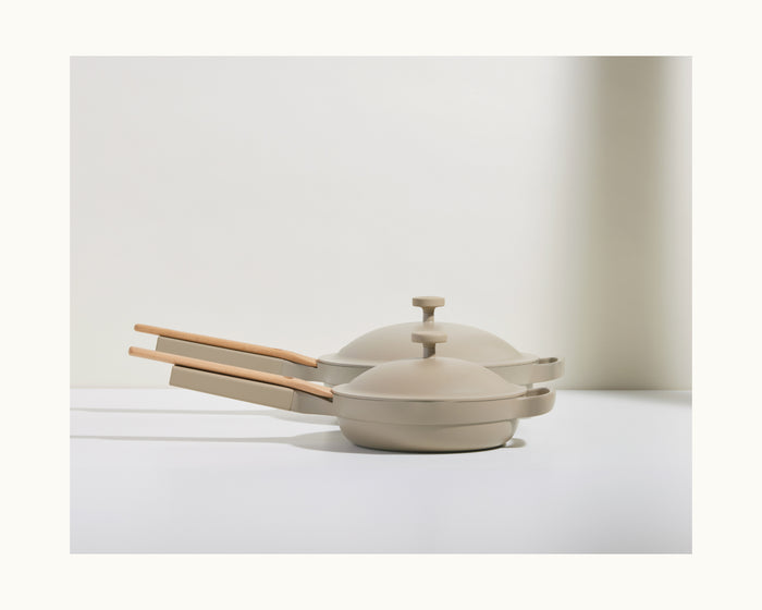 Two beige Mini Always Pan 2.0 sets with lids and wooden handles are stacked on a white surface, highlighting their multifunctional, non-toxic design for versatile cooking.