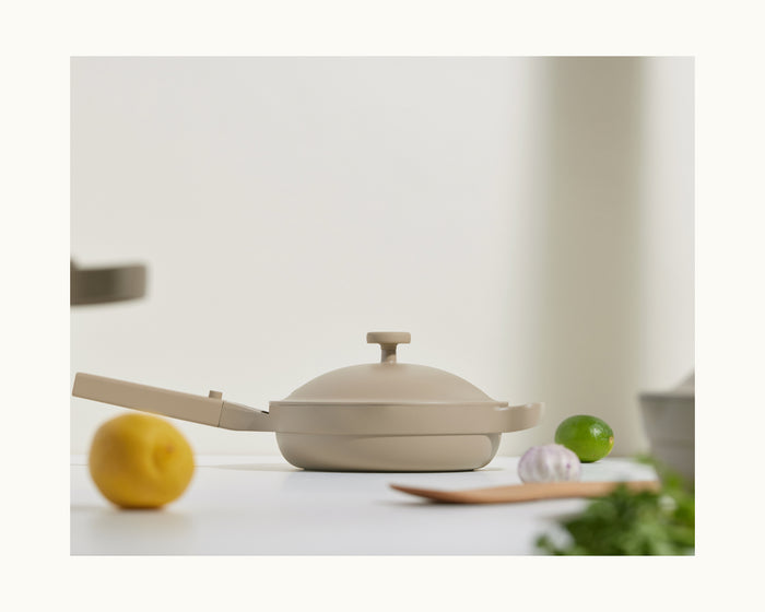 The Mini Always Pan 2.0 in beige, complete with a lid, rests on a white surface next to a lemon, lime, fresh herbs, and a wooden utensil.