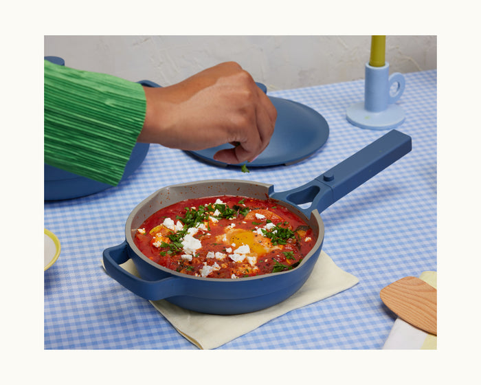 A hand sprinkles herbs over a skillet of tomato-based stew with eggs and cheese, all sizzling in the Mini Always Pan 2.0 atop a blue checkered tablecloth.