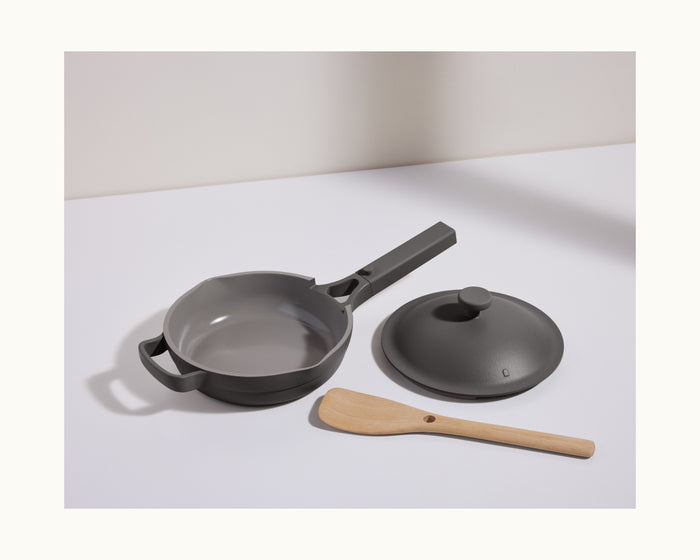 The Mini Always Pan 2.0 in gray, featuring a matching lid and wooden spatula, is displayed on a white surface with a shadow, highlighting this versatile and multifunctional kitchen pan.