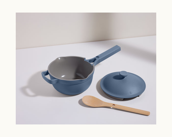 The Mini Perfect Pot 2.0, a blue nonstick pot with a matching lid, sits on a white surface beside a light wooden spoon.