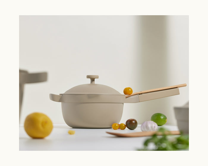 The Mini Perfect Pot 2.0 in beige, complete with lid and wooden spoon, rests on a white surface alongside a lemon, lime, tomatoes, garlic, and fresh herbs.