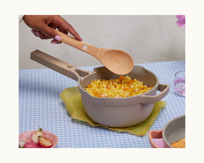 A hand holds a wooden spoon above the Mini Perfect Pot 2.0, which is filled with rice and vegetables, resting on a blue checkered tablecloth.