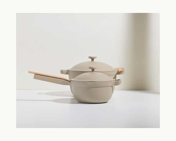 Two beige Mini Perfect Pot 2.0 pots with matching lids and light wooden handles are stacked on a white surface against a light background.