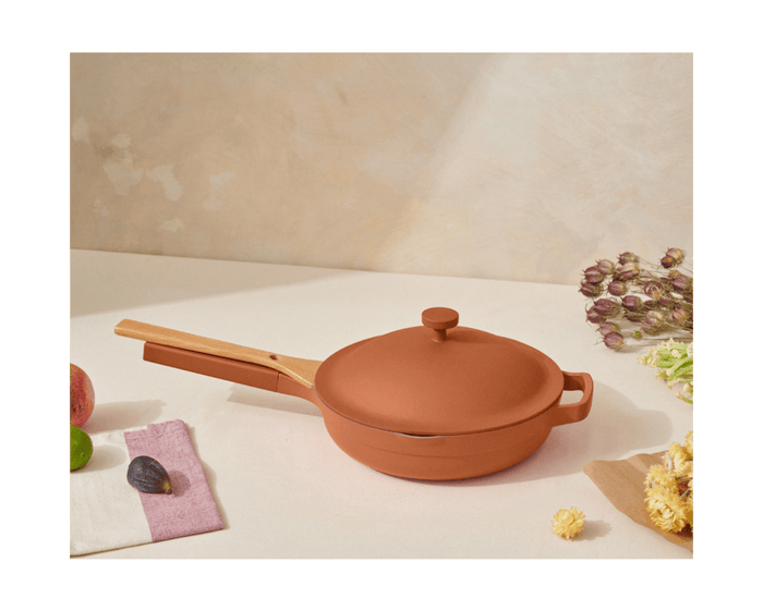 The Cookware Duo, a terracotta-hued pan with a lid and wooden handle from a bestselling set, rests on a light surface surrounded by fruits, a towel, and dried flowers.