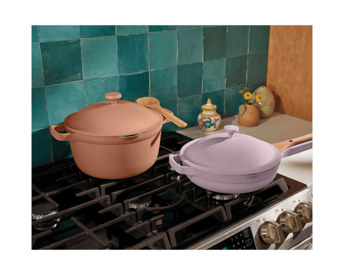 The Cookware Duo, featuring a brown Dutch oven and a lavender skillet, sits on a gas stove with kitchen utensils and decorative items in the background.