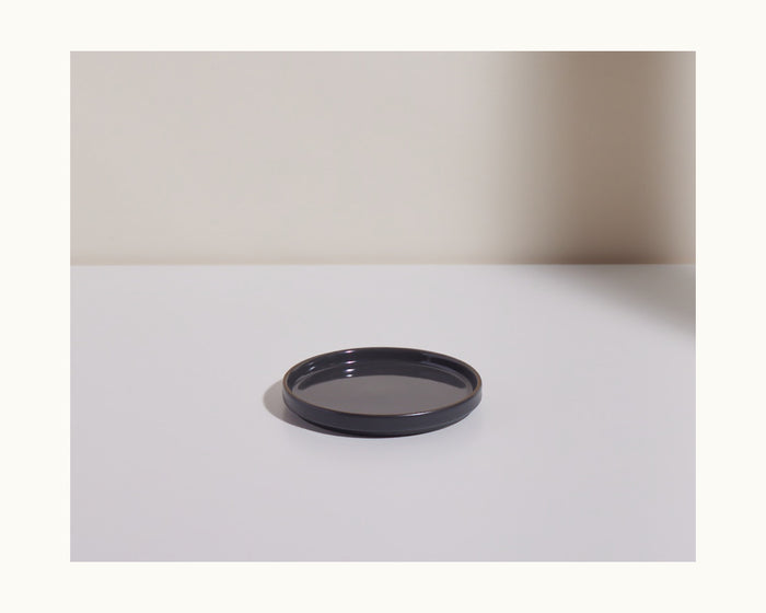 The Dessert Plates feature a round, black ceramic design and are shown on a white surface with a neutral background and a shadow on the right side.
