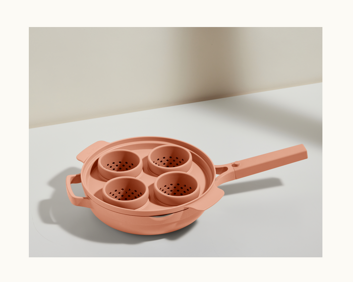 The Egg Poacher, a peach-colored pan with lid and four steamer baskets, is ideal for making poached eggs using silicone cups and looks elegant on a white surface.