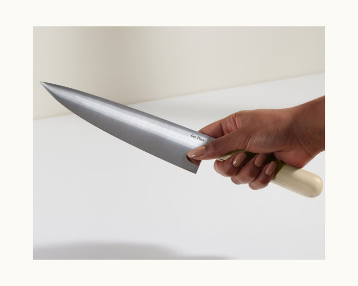 A hand holds the Everyday Chef’s Knife, featuring a large German steel blade and a light-colored handle, over a white surface.