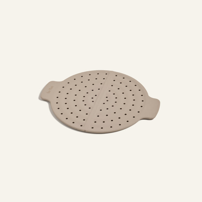 The Fearless Fry is a beige round silicone steamer insert with two side handles and multiple small holes, designed to fit perfectly in the Always Pan or to be used as a versatile frying tool.