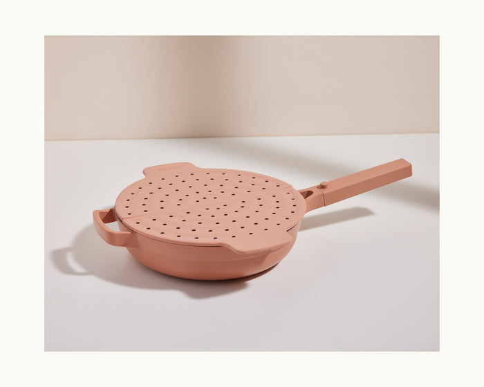 The Fearless Fry is a pink nonstick pan with two handles and a matching perforated splatter guard lid, shown on a white surface against a neutral background.