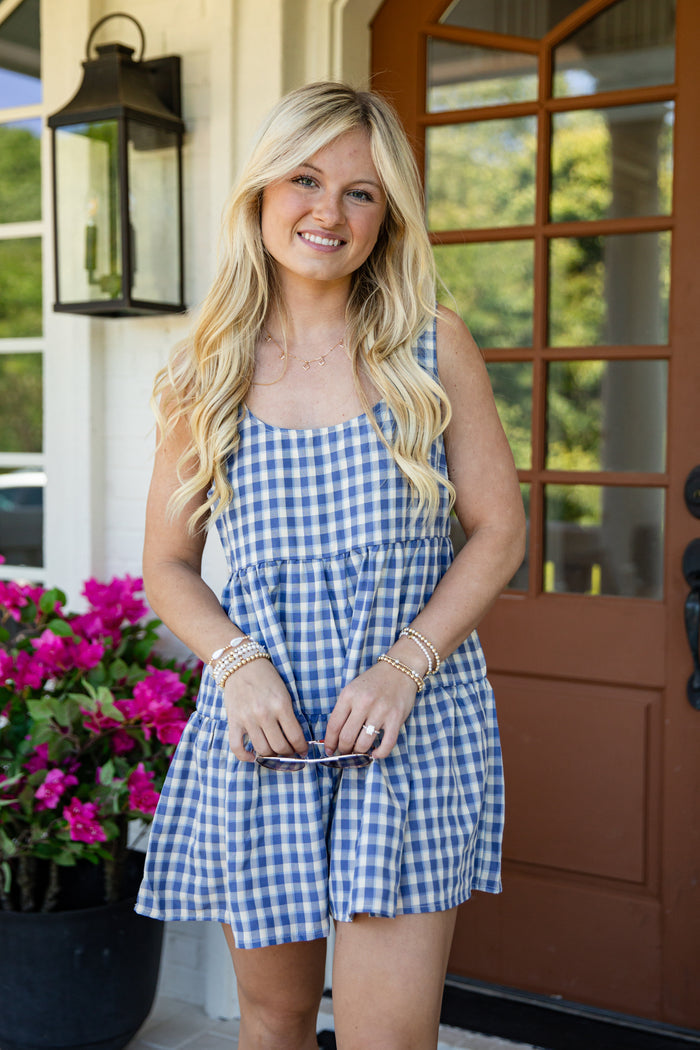 A smiling blonde woman wearing a blue and white gingham sleeveless romper stands on a front porch next to a brown door and pink flowers.