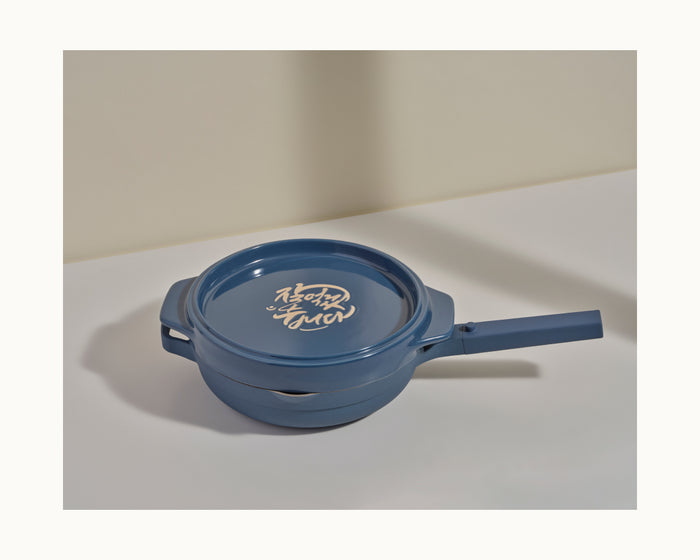 The Flipping Platter, a blue ceramic pot with a handle and script-embellished lid, rests on a white surface against a beige backdrop—ideal for serving tahdig or as an elegant flipping dish.