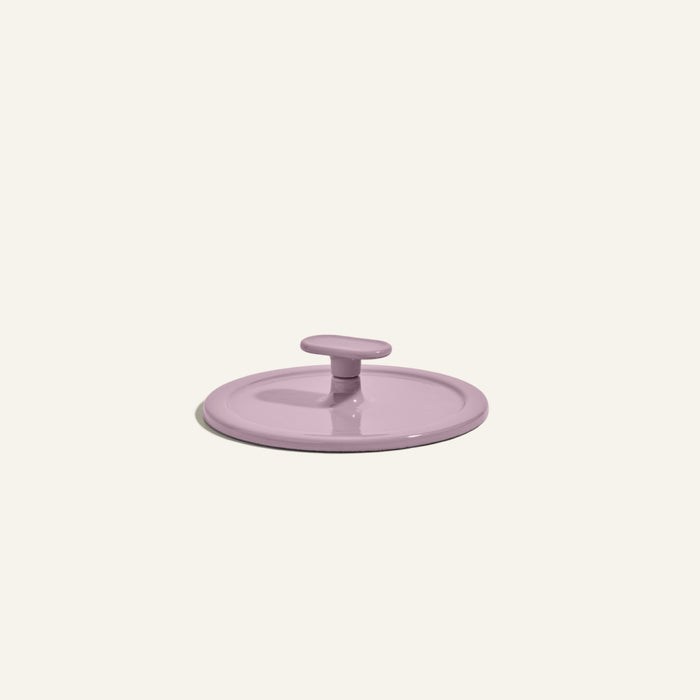 A round, light purple Grill Press with a central handle is shown against a plain off-white background, crafted from cast iron for even heat distribution.