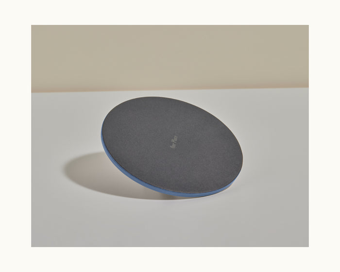 A Grill Press, round and black with a blue edge, sits on a white surface, its design echoing even heat distribution while casting a shadow to the left.