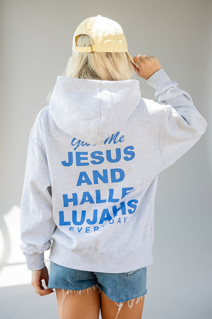 A person shown from behind wearing a light heather gray hoodie with bold blue text on the back reading 'You Me JESUS AND HALLE LUJAHS EVERY DAY,' paired with denim shorts and a cream baseball cap against a plain light gray background.