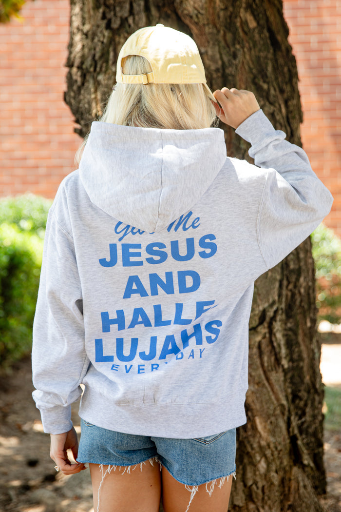 A woman viewed from behind wearing a light gray heather hoodie with bold blue text on the back reading 'Give Me JESUS AND HALLELUJAHS EVERY DAY.' She is standing outdoors next to a large tree trunk, wearing denim shorts and a gold baseball cap.