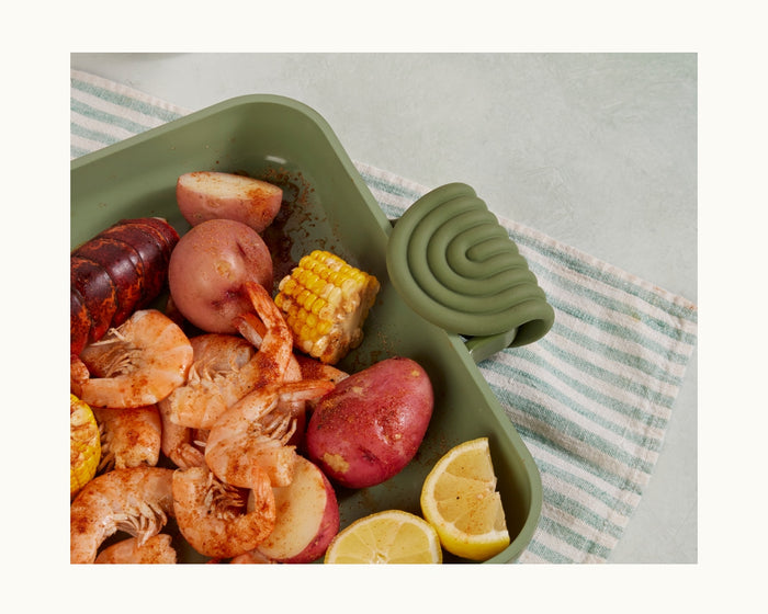 Hot Grips, a green baking dish with helper handles, is filled with shrimp, red potatoes, corn on the cob, lemon wedges, and sausage—ideal for easy carrying and kitchen protection.