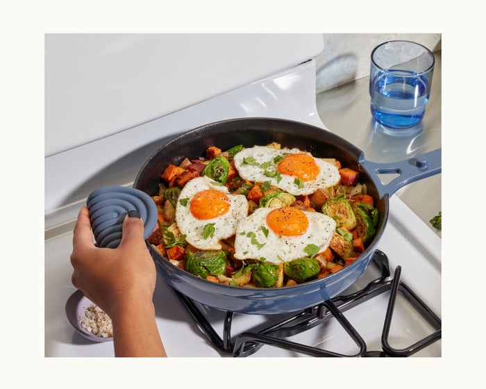 A hand uses Hot Grips to hold a pan with helper handles on a stove, cooking roasted vegetables and two sunny-side-up eggs, with a glass of water and a small bowl nearby for added kitchen safety.
