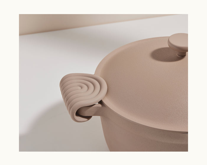 A close-up of a beige pot with a lid, featuring the spiral-patterned Hot Grips silicone handle cover for kitchen protection, shown on a light surface.