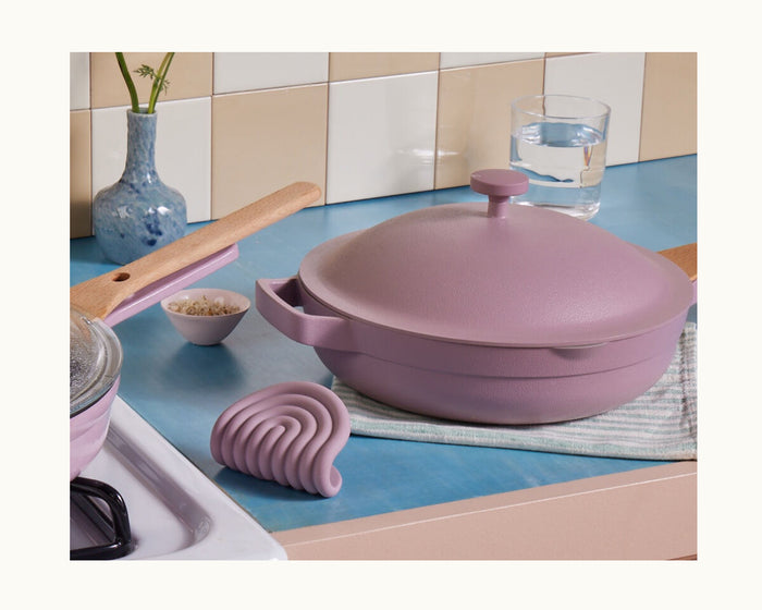 A lavender Hot Grips pot with helper handles and a lid sits on a blue countertop beside a matching pan, silicone utensil rest, small bowl of grains, flower vase, and glass of water.