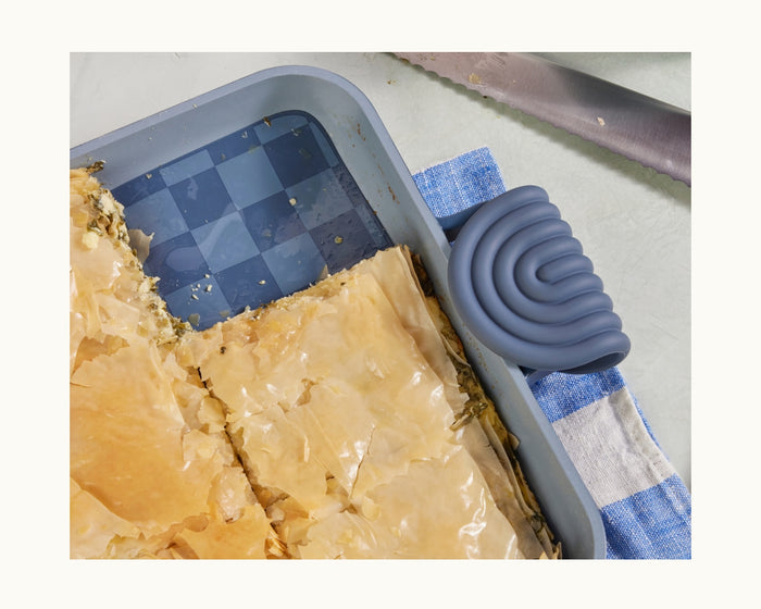 The Hot Grips rectangular pan, featuring helper handles for easy lifting, holds pieces of baked pastry and sits on a blue and white towel next to a serrated knife on a light-colored surface.