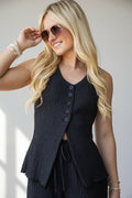 A woman wearing a sleeveless black textured knit vest with a button-front closure and drawstring waist, styled with sunglasses and bracelets against a light gray background.