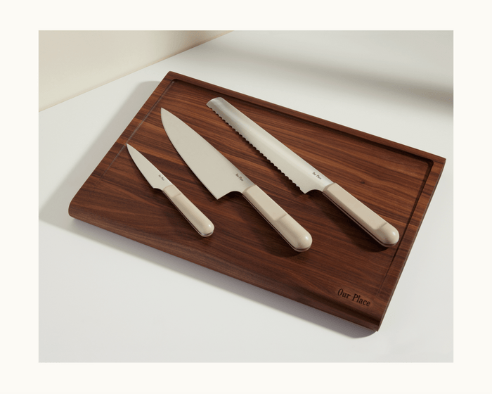 The Knife Trio, featuring beige-handled serrated slicing, paring, and chef’s knives, is arranged on a dark wooden Our Place cutting board against a light background.