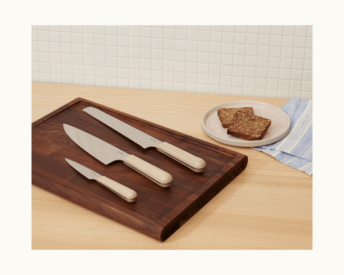 The Knife Trio, featuring beige-handled Serrated Slicing, Paring, and Chef’s Knives, is arranged on a wooden cutting board beside a plate with two bread slices on a light wood countertop.