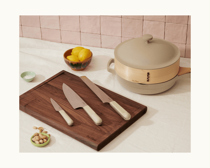 A Knife Trio set with a cutting board, a bowl of lemons, a bowl of pistachios, a small dish of salt, and a beige pot with lid displayed on a white tablecloth.