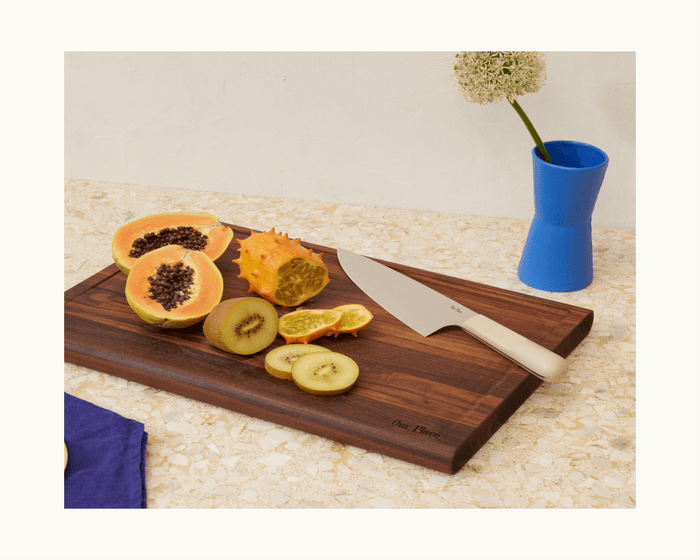 A Knife Trio and a cutting board with sliced papaya, kiwifruit, horned melon, and tangerine rest on a countertop beside a blue vase holding a white flower.