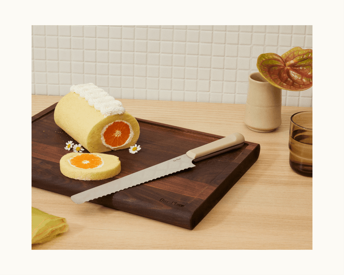 A sliced rolled cake with an orange pattern sits on a wooden board beside the Knife Trio, a cup, a vase with a plant, and small flowers on a light wood surface.