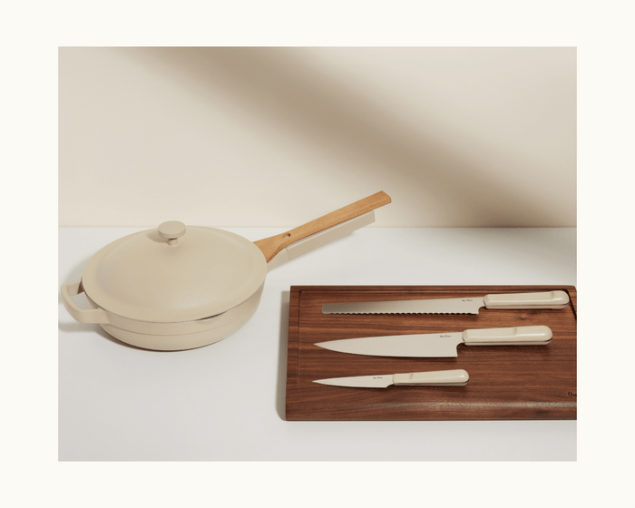 A cream-colored pan with a lid and wooden handle sits next to a wooden cutting board holding the Knife Trio—Chef’s Knife, Paring Knife, and Serrated Slicing Knife—all featuring matching cream handles.