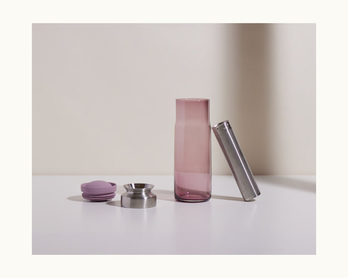 The Night + Day Carafe set includes a tall pink glass cold brew infuser bottle, metal jigger, metal strainer, and two purple silicone accessories, all displayed on a white surface against a neutral background.