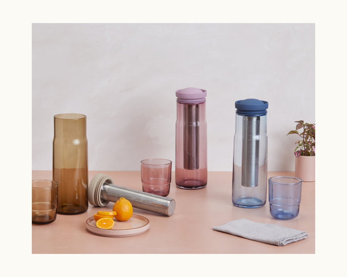 Three Night + Day Carafes with colored lids and metal infusers are arranged alongside matching cups, a napkin, cut citrus, and a small potted plant on a pastel surface.