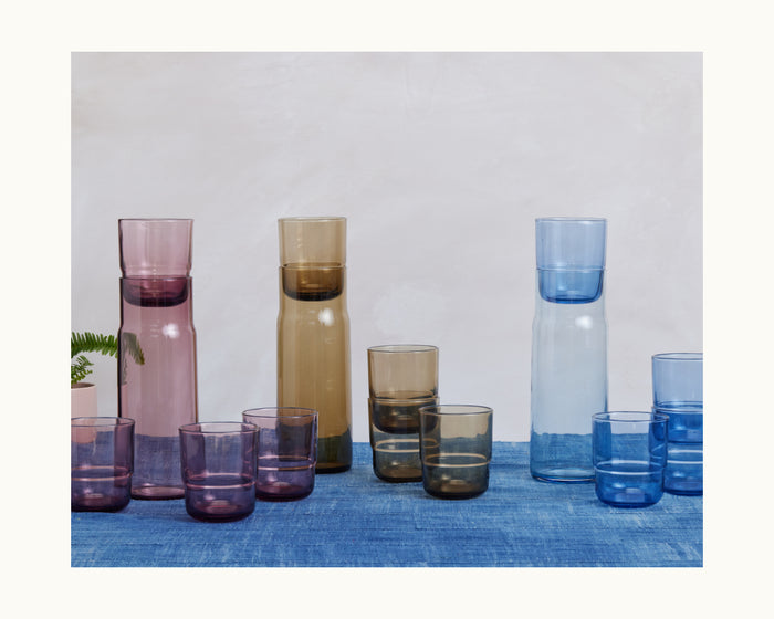 Three Night + Day Carafe sets in purple, brown, and blue—each with matching tumblers—are arranged on a blue tablecloth against a light background, ideal for serving drinks or as a stylish wine aerator.