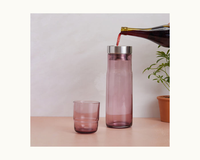 The Night + Day Carafe, tinted pink, is being filled with red wine from an aerator beside its matching glass and a potted plant on a light surface.