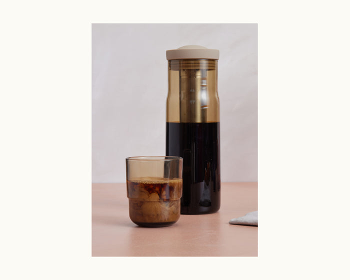 The Night + Day Carafe, filled with dark cold brew, stands beside a small glass of creamy iced coffee on a light surface.
