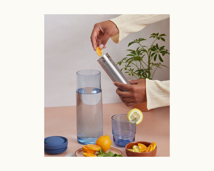 Someone adds a lemon wedge to the stainless steel infuser of the Night + Day Carafe above a glass of water; on the table are a pitcher, sliced lemons, and blue lids.