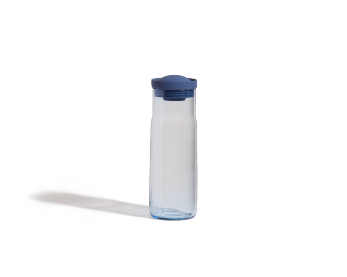 The Night + Day Carafe, a transparent plastic water bottle with a blue lid and sleek carafe design, stands upright on a white background, casting a faint shadow.